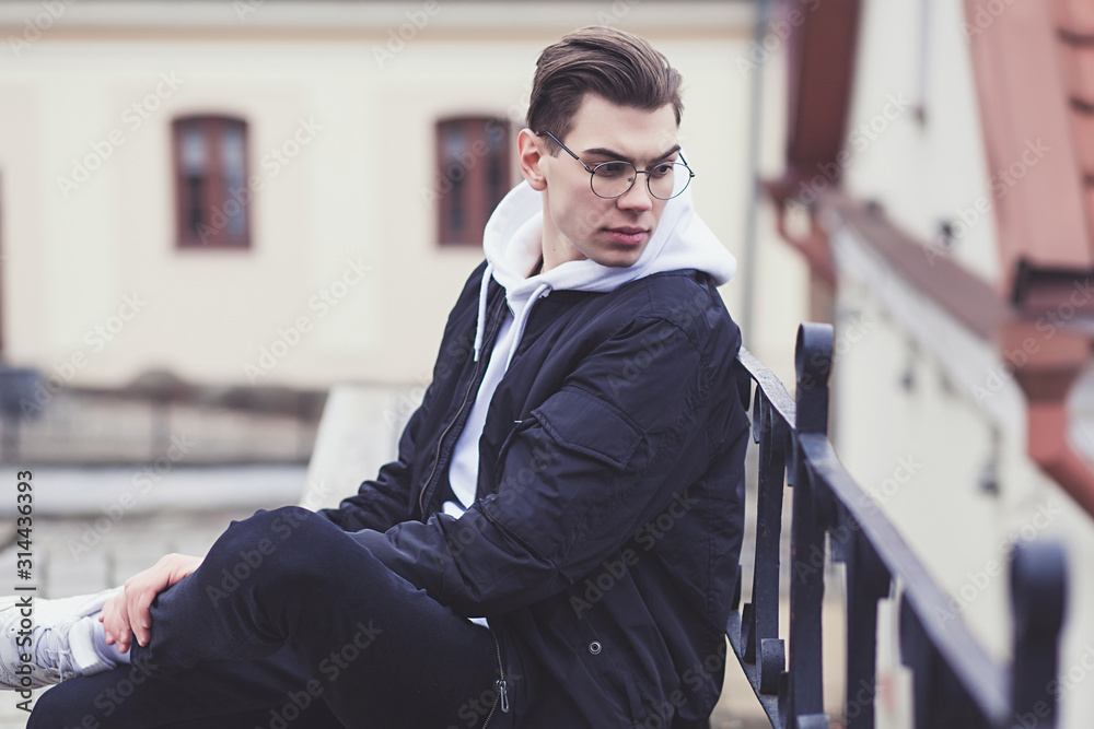 young man in a black autumn jacket and sneakers sits on the street young man in a black autumn jacket and sneakers sits on the street