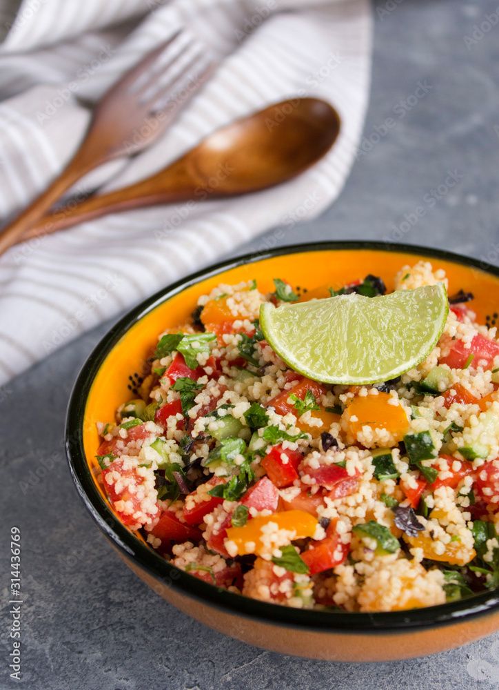 Tabouleh: vegetable couscous salad. Vegetables and couscous salad tabouleh in the orange ceramic bowl on concrete surface. Fresh summer salad with lime slice, wooden spoon, fork and white linen towel Tabouleh: vegetable couscous salad. Vegetables and couscous salad tabouleh in the orange ceramic bowl on concrete surface. Fresh summer salad with lime slice, wooden spoon, fork and white linen towel