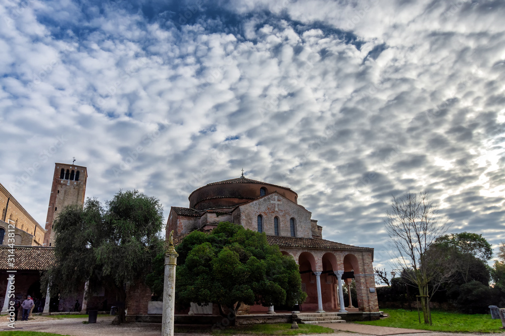 View of Torcello Cathedral (Basilica di Santa Maria Assunta), Church of Santa Fosca and the bell tower, over green trees under blue sky, on the island of Torcello