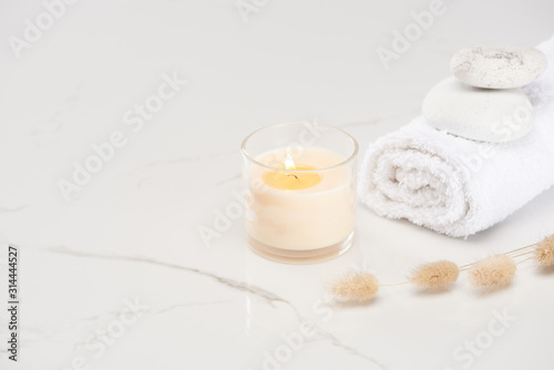 fluffy bunny tail grass near burning white candle in glass and rolled towel with stones on marble white surface