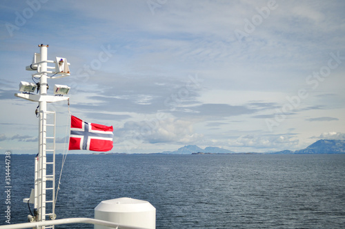 View to the flag of Norway on ferry deck on windy day with Vega archipelago mountains in the background in Nordland county, Norway on windy and cloudy summer day