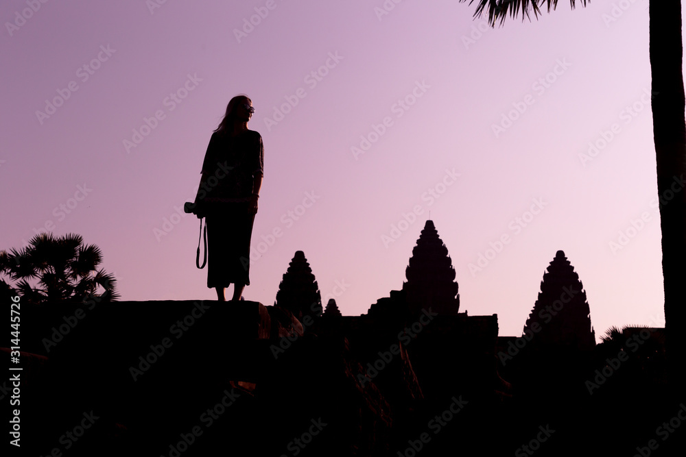 Silhouette of Angkor Wat and girl traveler with photo camera on the sunrise. Angkor Wat - Hindu temple complex in Cambodia, largest religious monument in the world. Popular tourist attraction.