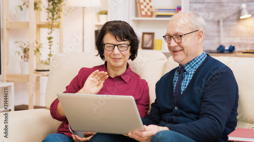 Elderly age couple sitting on sofa holding laptop during a video call.