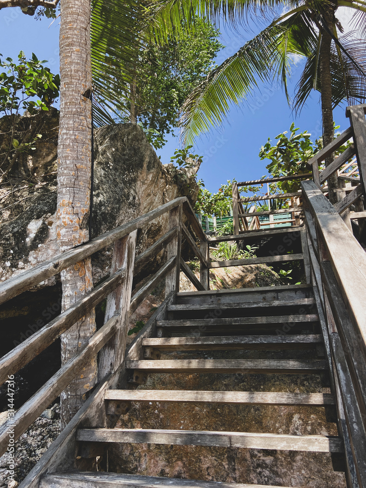 An old wooden staircase in the tropics on an island on the coast among green palm trees and a blue sky. Steps leading up. Trees next to large stones on a beautiful beach.