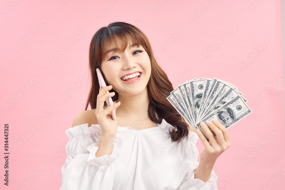 Portrait of a cheerful young woman holding money banknotes and celebrating isolated over pink background. Using mobile phone. Portrait of a cheerful young woman holding money banknotes and celebrating isolated over pink background. Using mobile phone.