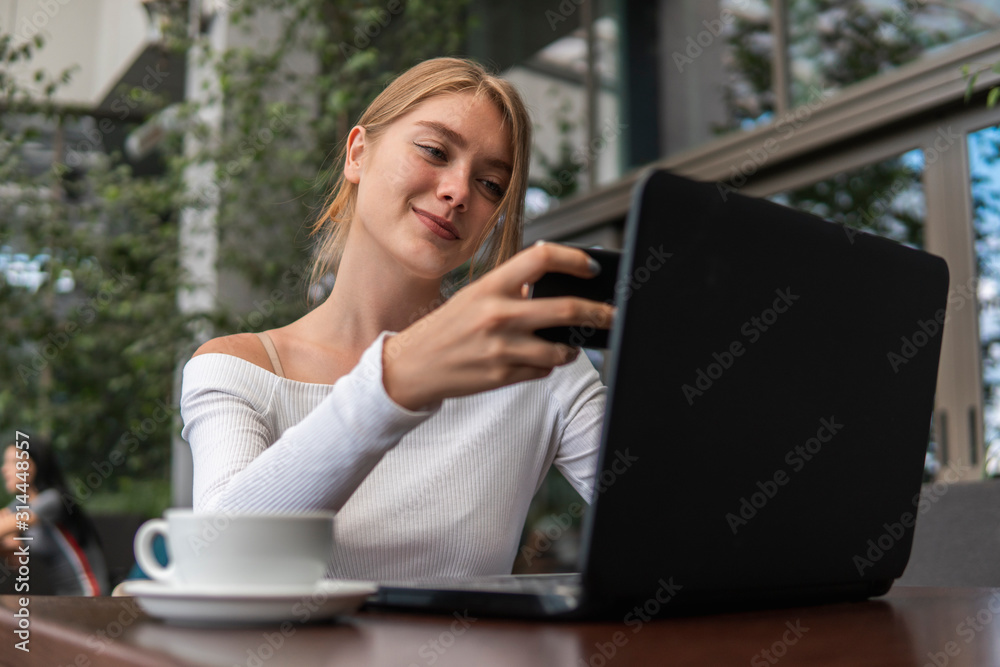 Modern business woman is reading incoming sms message on smartphone or watching broadcasting online on modern mobile phone while sitting in caffe with a laptop. Female student sitting in coffee. Modern business woman is reading incoming sms message on smartphone or watching broadcasting online on modern mobile phone while sitting in caffe with a laptop. Female student sitting in coffee.