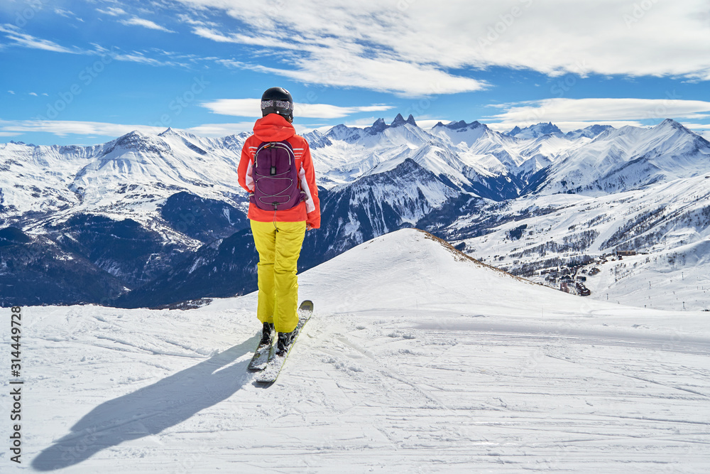 Female skier with colorful clothes looking at the Les Aiguilles d'Arves peaks, from a ski slope high above the Les Sybelles ski domain, in France, on a sunny Winter day. Ski holiday concept.