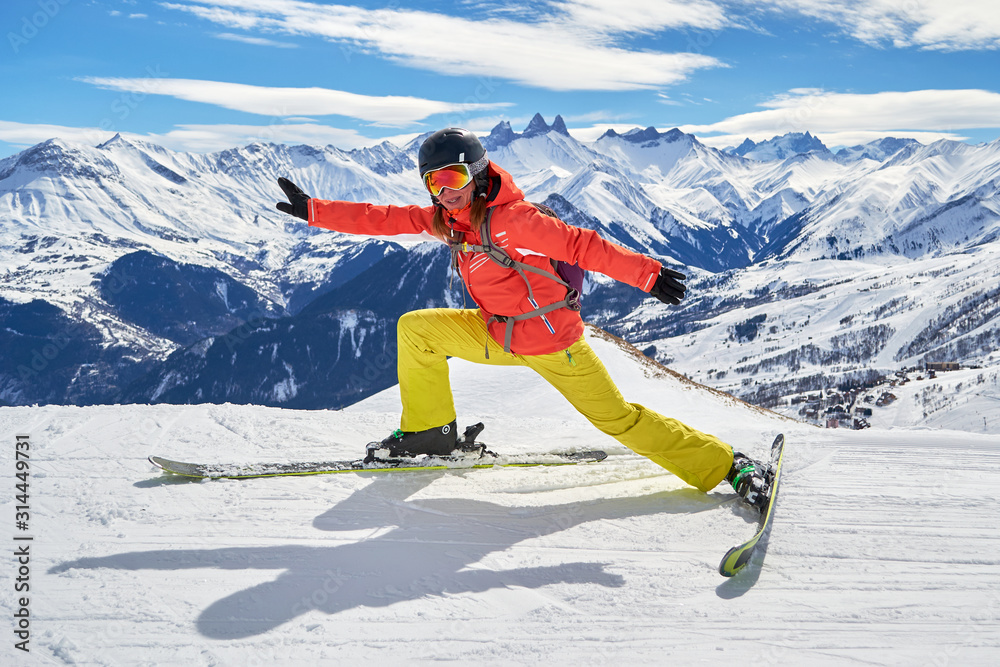 Woman skier stretching on skis with a funny pose, high above on a ski piste in Les Sybelles ski domain, France, on a sunny Winter day, with Les Aiguilles d'Arves peaks in the background.