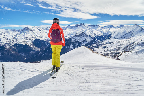 Female skier with colorful clothes looking at the Les Aiguilles d'Arves peaks, from a ski slope high above the Les Sybelles ski domain, in France, on a sunny Winter day. Ski holiday concept.