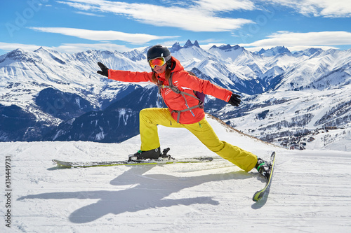 Obraz na płótnie Woman skier stretching on skis with a funny pose, high above on a ski piste in Les Sybelles ski domain, France, on a sunny Winter day, with Les Aiguilles d'Arves peaks in the background. w salonie