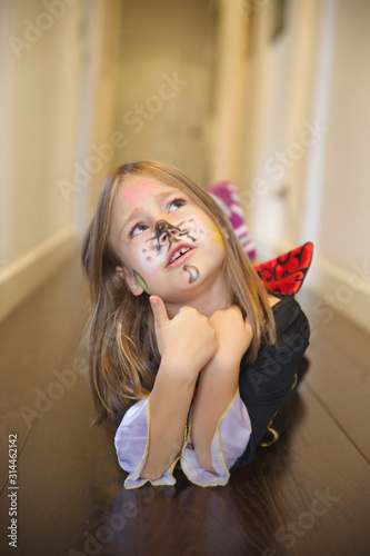 Portrait of blonde six years old girl disguised for Halloween with painted face with thinking expression, enumerating with her fingers, lying in corridor inside home