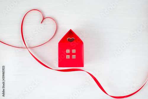 Red ribbon heart on a white wooden background, red house with a heart shape window in a romantic, valentine, family or wedding concept. Empty space for your text.