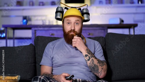 Bearded man with beea helmet sitting on the sofa at home, drinking beer, watching tv and relaxing. photo