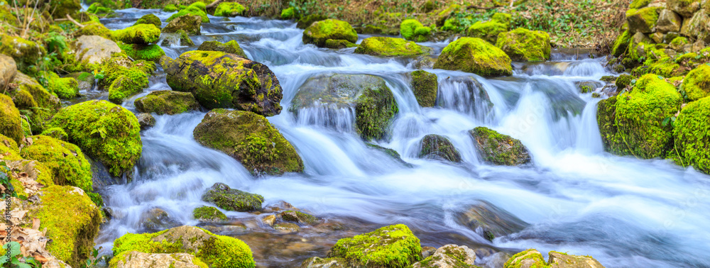 A small stream with a waterfall and mossy rocks in spring, panoramic image - obrazy, fototapety, plakaty 
