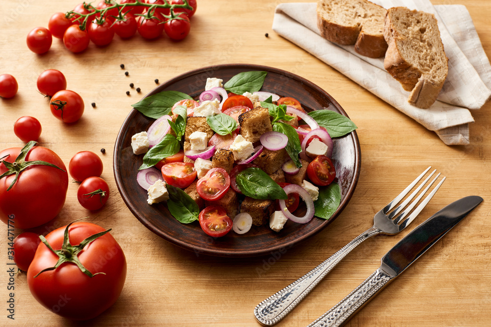 delicious Italian vegetable salad panzanella served on plate on wooden table near fresh tomatoes, bread, fork and knife