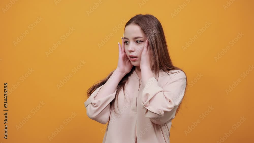 Surprised caucasian brunette woman in fashion pastel shirt keeping palms on cheeks, opened mouth, in shock on orange background in studio. People sincere emotions, lifestyle concept. Surprised caucasian brunette woman in fashion pastel shirt keeping palms on cheeks, opened mouth, in shock on orange background in studio. People sincere emotions, lifestyle concept.