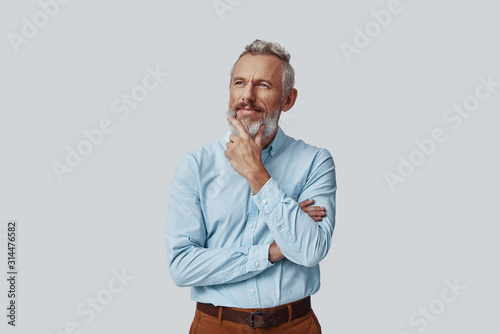 Thoughtful mature man looking at camera and keeping hand on chin while standing against grey background