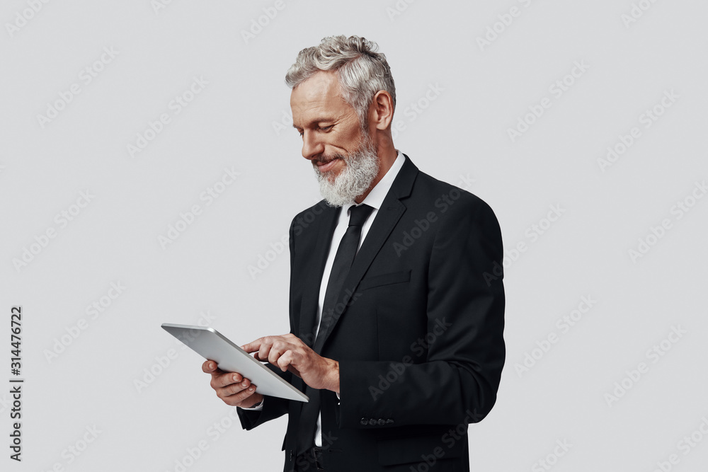 Thoughtful mature man in full suit working using digital tablet and smiling while standing against grey background Thoughtful mature man in full suit working using digital tablet and smiling while standing against grey background