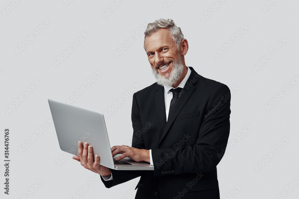 Confident mature man in full suit working using laptop and smiling while standing against grey background Confident mature man in full suit working using laptop and smiling while standing against grey background