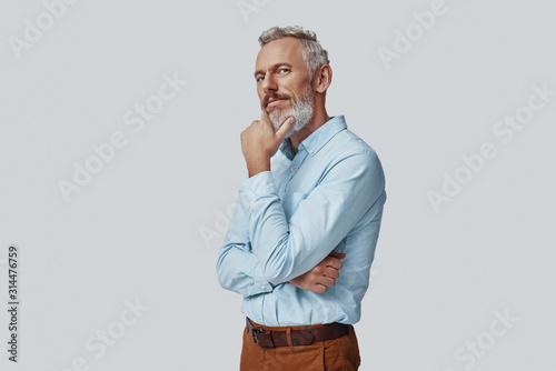 Thoughtful mature man looking at camera and keeping hand on chin while standing against grey background
