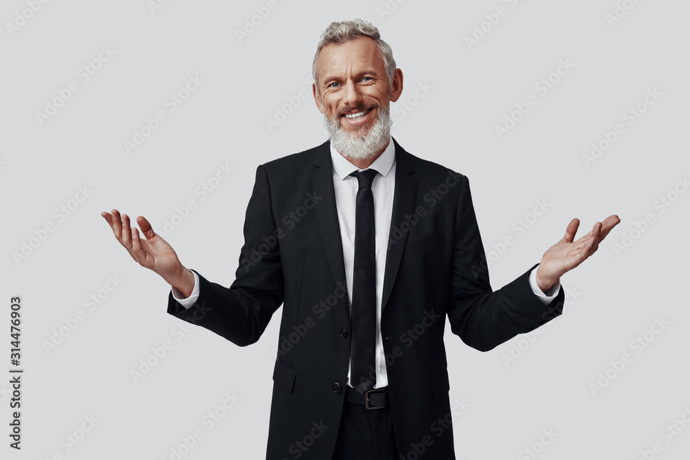 Confident mature man in full suit keeping arms outstretched and looking at camera while standing against grey background Confident mature man in full suit keeping arms outstretched and looking at camera while standing against grey background