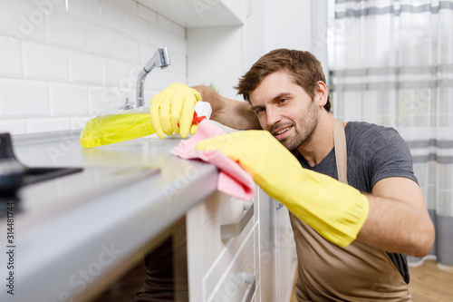 Close-up of young man in yellow gloves using cleanser to wash kitchen furniture