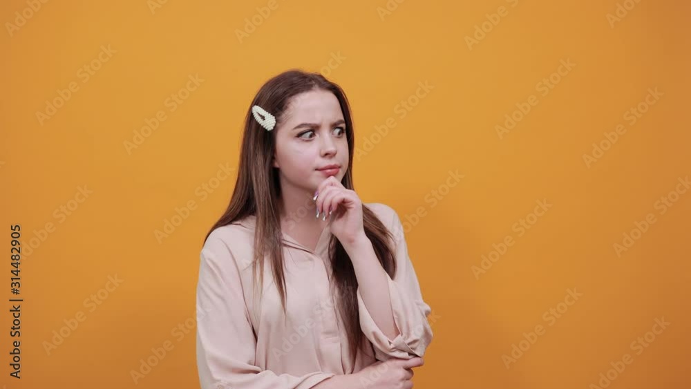 Disappointed woman in pastel shirt keeping fins over chin, holding other hand on stomach, thinking about issue isolated on orange background in studio. People sincere emotions, lifestyle concept. Disappointed woman in pastel shirt keeping fins over chin, holding other hand on stomach, thinking about issue isolated on orange background in studio. People sincere emotions, lifestyle concept.