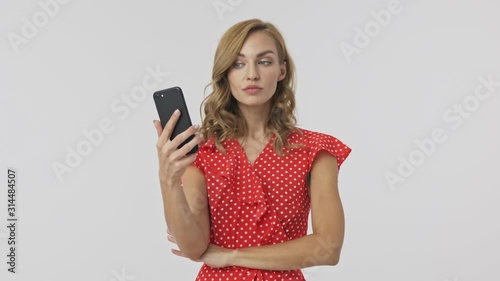 Calm pretty blonde woman in dress using smartphone like a mirror and smartening over grey background photo