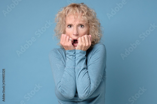 Nervous mature woman bites fingernails, feel anxious, afraids of doctors, has scared expression, worries someone will know her secret. Studio shot
