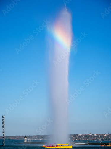 A rainbow appears in the spray across the iconic water fountain located in Geneva, Switzerland. A tourist boat passes below the fountain on the lake front.