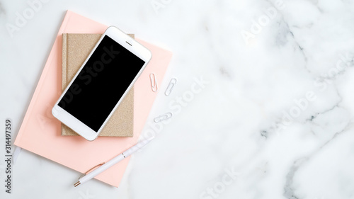 Home office desk workspace with smartphone screen mockup, pink notepad, pen on marble background. Flat lay, top view. Elegant feminine workplace with stylish accessories