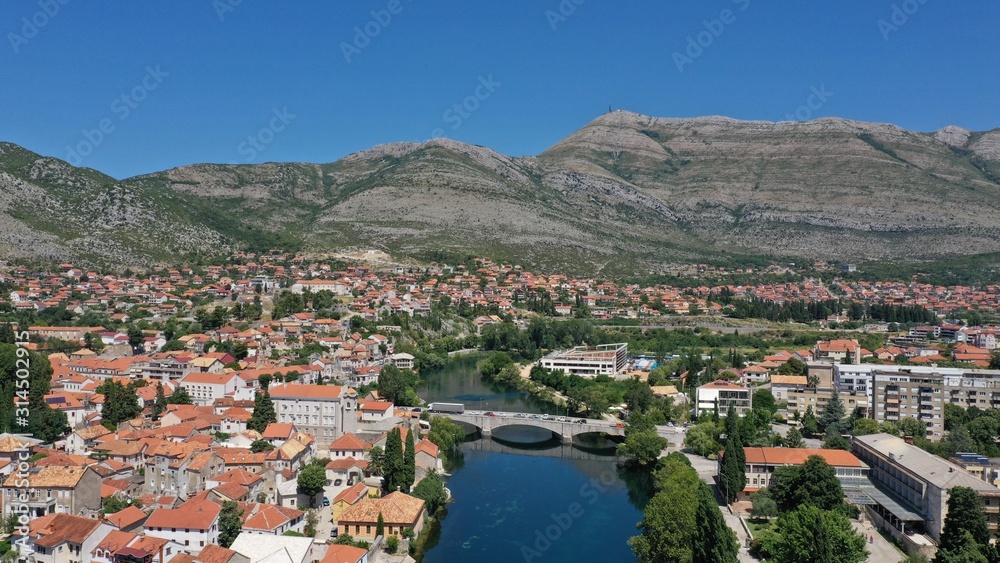 Aerial view of stone bridge (Kameni Most) on Trebisnjica river in Trebinje Old Town. Bosnia and Herzegovina. Summer sunny day, Turquoise water, mountains, trees, blue sky, small houses.