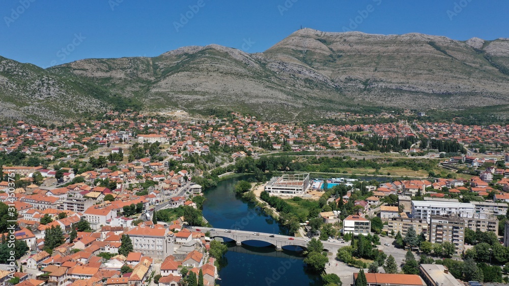 Naklejka premium Aerial view of stone bridge (Kameni Most) on Trebisnjica river in Trebinje Old Town. Bosnia and Herzegovina. Summer sunny day, Turquoise water, mountains, trees, blue sky, small houses.