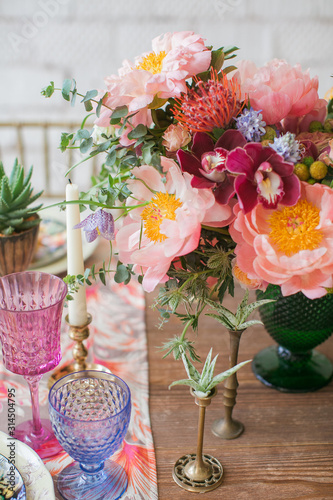 Served table for a wedding dinner in boho chic style. Decor from fresh flowers of peonies, roses, orchids and Leucospermum conocarpodendron. Crockery plates, glasses and candles. Wooden table.