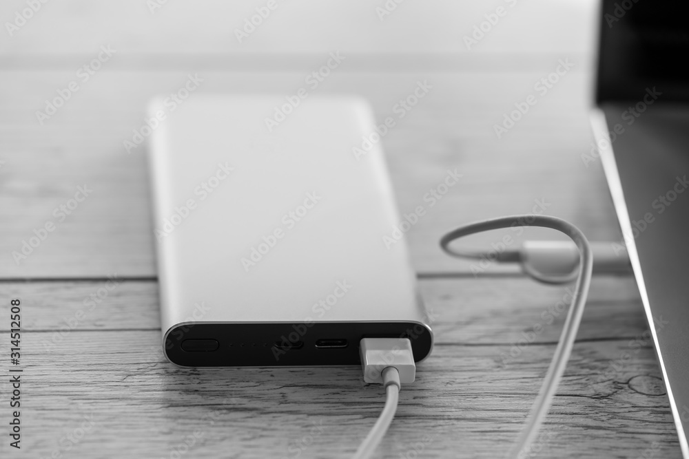 Laptop charging from silver power bank, charger battery on wooden background. Productive portable device for business traveling, black and white image, high angle view and macro Laptop charging from silver power bank, charger battery on wooden background. Productive portable device for business traveling, black and white image, high angle view and macro