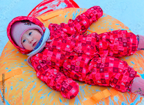 Cute Asian little girl a pink colorful jumpsuit portrait smiling sliding on orange tube on a white fluffy snow . Happy baby outdoors in snow on cold winter day.