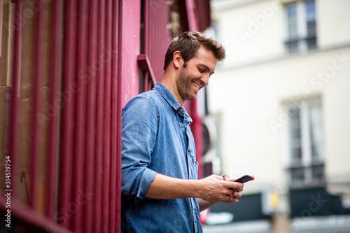Side of happy man leaning against wall looking at mobile phone in city