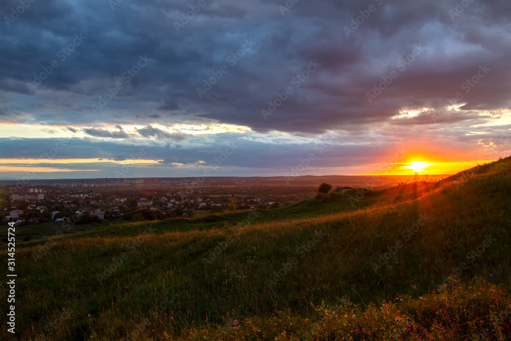 Sunset in dramatic cloudscape over wild herb field with distant city in view.