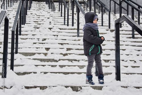 A little boy stands on a snow-covered staircase in the street in winter. © Konstiantyn Zapylaie