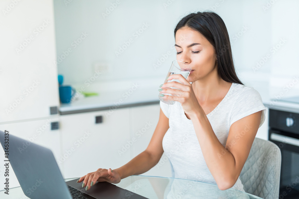 Young Woman sitting in kitchen table, looking at laptop computer screen while drink water from glass Young Woman sitting in kitchen table, looking at laptop computer screen while drink water from glass