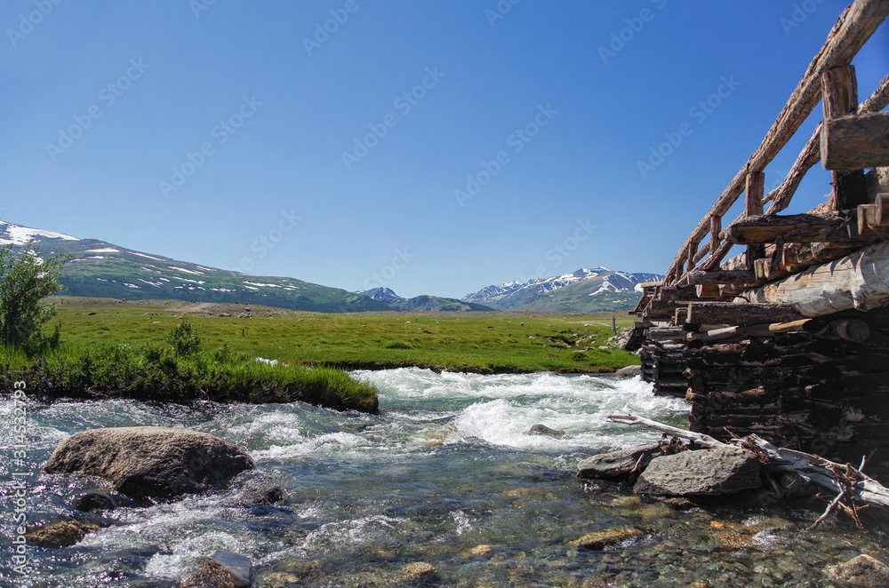 Mongolian Altai. Log bridge through the raging mountain river. High water. Spring run-off. Nature and travel. Mongolia, Bayan-Olgii Province, Altai Tavan Bogd National Park Mongolian Altai. Log bridge through the raging mountain river. High water. Spring run-off. Nature and travel. Mongolia, Bayan-Olgii Province, Altai Tavan Bogd National Park