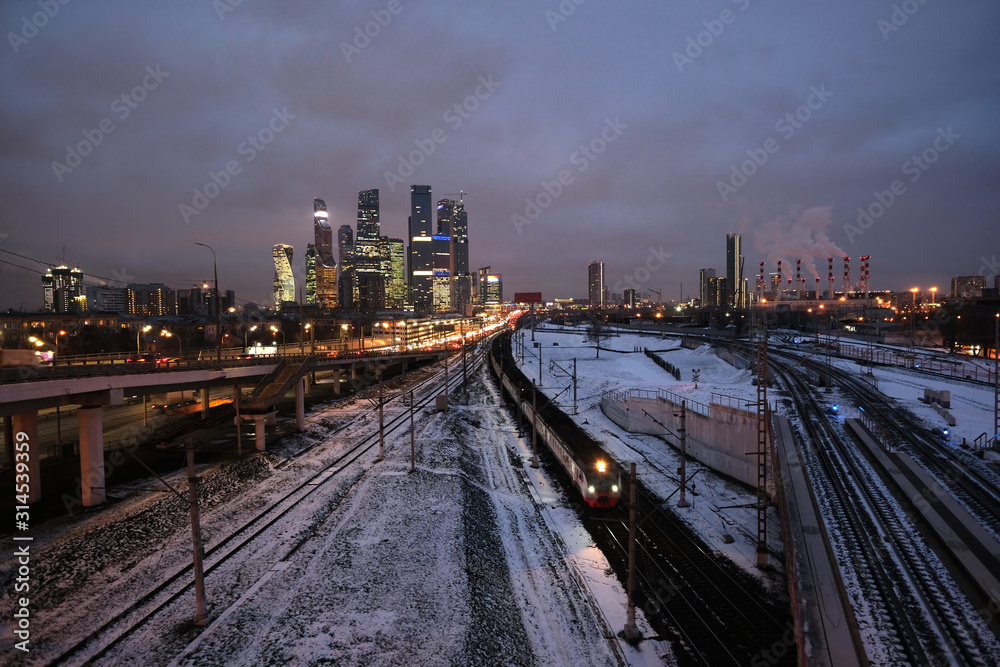Industrial landscape with many railroad tracks going far, cars on the overpass and skyscrapers with power plant pipes on horizon on twilight in winter season