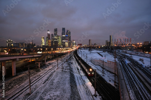 Industrial landscape with many railroad tracks going far, cars on the overpass and skyscrapers with power plant pipes on horizon on twilight in winter season