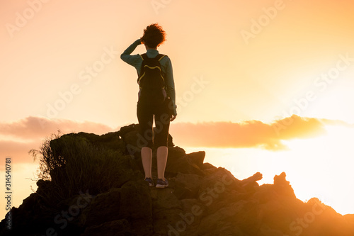 Back view of a sporty trekker girl standing on the rocks, looking at the horizon. Woman with backpack walking in the evening at sunset. Backlight silhouette. Sport adventure freedom and hiking concept