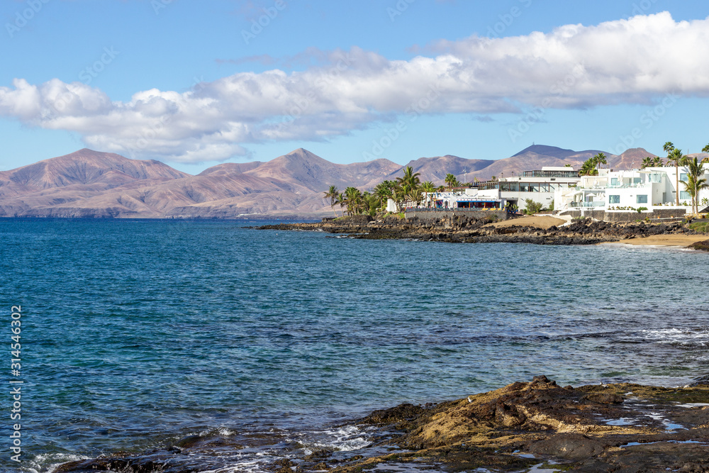 Rocky coast of Puerto del Carmen at Canary island Lanzarote with lava rocks and blue water in the foreground, white houses, palm trees and volcanic mountain range in the background