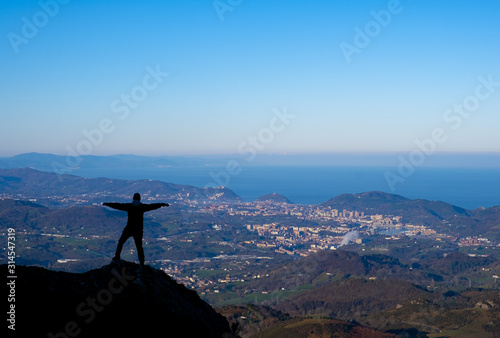 Mountaineer with arms raised on top of Aiako Harriak with the city of Donostia in the background, Euskadi