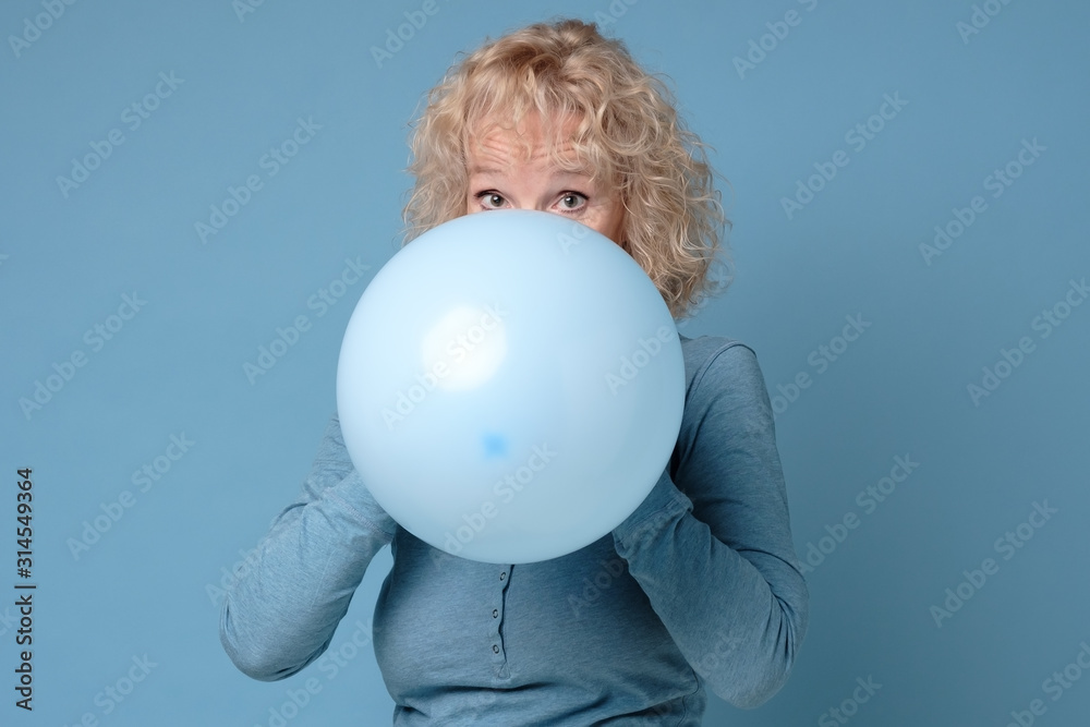 Beautiful senior blonde woman preparing for party blowing up blue balloon Beautiful senior blonde woman preparing for party blowing up blue balloon