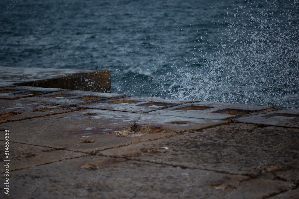 A sea wave hits the pier from stone slabs, water splashes flying to the sides. Nobody is on the waterfront. Seascape with storm surge and sea spray. Black Sea, Odessa, Ukraine. A sea wave hits the pier from stone slabs, water splashes flying to the sides. Nobody is on the waterfront. Seascape with storm surge and sea spray. Black Sea, Odessa, Ukraine.