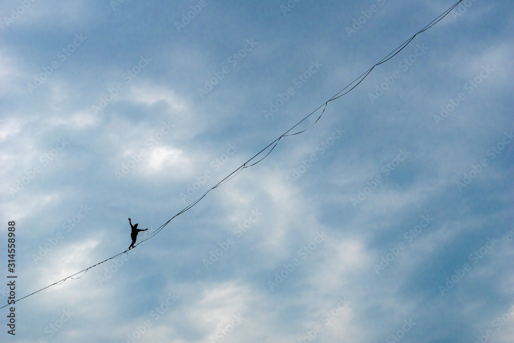 A man walking on strecthed sling - performance of a tightrope walker with background of dramatic clouds. Man balances over the abyss taking a  decisive step.
