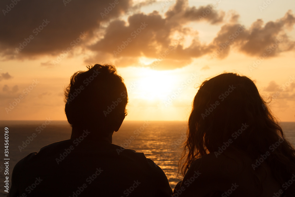 A guy and a girl meet the sunset while sitting on a mountain. Silhouettes of a couple in love who are sitting with their backs to the camera and watching the bright sunset over the ocean. Romance,love